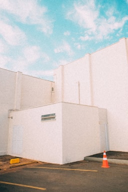 A minimalistic architectural scene featuring white building structures under a bright blue sky. The geometric shapes of the buildings create a clean and modern aesthetic. A single orange traffic cone is placed on the ground near the corner of the building, adding a pop of color.