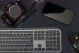 A flat lay featuring a camera, smartphone, keyboard, and computer mouse, all arranged on a dark surface. Two pens, including a stylus, are also present. The layout suggests a workspace with electronic devices and accessories placed closely.
