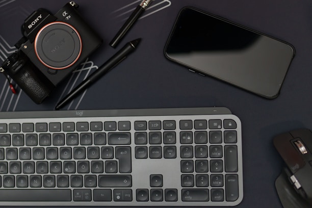 A flat lay featuring a camera, smartphone, keyboard, and computer mouse, all arranged on a dark surface. Two pens, including a stylus, are also present. The layout suggests a workspace with electronic devices and accessories placed closely.