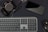 A flat lay featuring a camera, smartphone, keyboard, and computer mouse, all arranged on a dark surface. Two pens, including a stylus, are also present. The layout suggests a workspace with electronic devices and accessories placed closely.