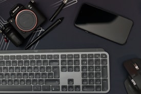A flat lay featuring a camera, smartphone, keyboard, and computer mouse, all arranged on a dark surface. Two pens, including a stylus, are also present. The layout suggests a workspace with electronic devices and accessories placed closely.