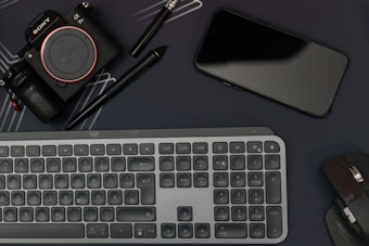 A flat lay featuring a camera, smartphone, keyboard, and computer mouse, all arranged on a dark surface. Two pens, including a stylus, are also present. The layout suggests a workspace with electronic devices and accessories placed closely.