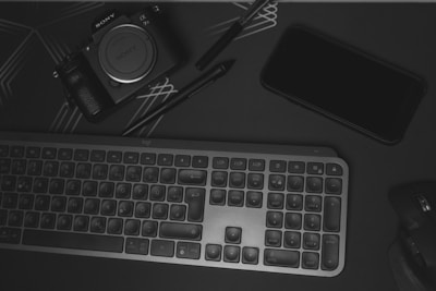 A flat lay of electronic devices and accessories on a dark surface. There is a Sony alpha series camera, a black smartphone, a compact keyboard, and a wireless mouse. A stylus or pen is also visible, and the surface has subtle geometric line patterns.