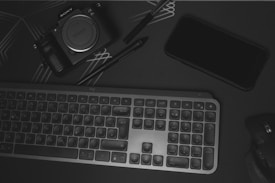 A flat lay of electronic devices and accessories on a dark surface. There is a Sony alpha series camera, a black smartphone, a compact keyboard, and a wireless mouse. A stylus or pen is also visible, and the surface has subtle geometric line patterns.