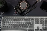 A workspace setup featuring a Sony camera, a computer keyboard, two pens, a smartphone, and a camera lens, all positioned on a dark surface with subtle line patterns.