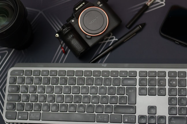 A workspace setup featuring a Sony camera, a computer keyboard, two pens, a smartphone, and a camera lens, all positioned on a dark surface with subtle line patterns.