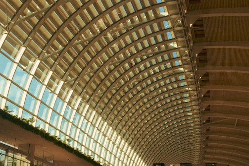 An expansive arched ceiling made of glass and metal beams allows natural light to flood into the space. The structure features repetitive geometric patterns in the form of curved beams and large glass panels. Greenery is visible along the lower portion, adding a touch of nature to the architectural setting.