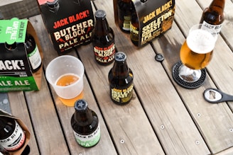 A variety of craft beer bottles and packs, including Jack Black's Butcher Block Pale Ale and Skeleton Coast IPA, are arranged on a wooden table. Some of the beers are in bottles while others are in cardboard carriers. A half-filled plastic cup and a glass of beer with a frothy head are also visible. A bottle opener rests on the table.