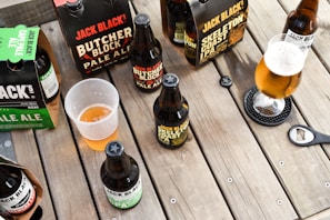 A variety of craft beer bottles and packs, including Jack Black's Butcher Block Pale Ale and Skeleton Coast IPA, are arranged on a wooden table. Some of the beers are in bottles while others are in cardboard carriers. A half-filled plastic cup and a glass of beer with a frothy head are also visible. A bottle opener rests on the table.
