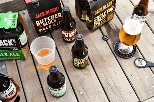 A variety of craft beer bottles and packs, including Jack Black's Butcher Block Pale Ale and Skeleton Coast IPA, are arranged on a wooden table. Some of the beers are in bottles while others are in cardboard carriers. A half-filled plastic cup and a glass of beer with a frothy head are also visible. A bottle opener rests on the table.