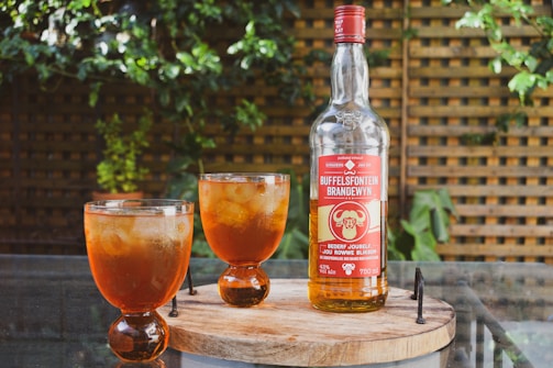 A bottle of Buffelsfontein Brandewyn is placed on a wooden round tray along with two glasses filled with an amber liquid and ice. The setup is outdoors, with green foliage and a lattice fence in the background, creating a rustic and natural atmosphere.