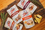 Five packets of Spur heat and pour sauces are arranged on a wooden surface. The flavors include Monkey Gland, Cheddamelt, Mushroom Sauce, and Pepper Sauce. Fresh sprigs of rosemary and thyme are placed beside the pouches, and there are chunks of cheese next to the Cheddamelt packet. The packaging is visually appealing, featuring images of the respective dishes the sauces can be used on.