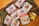 Five packets of Spur heat and pour sauces are arranged on a wooden surface. The flavors include Monkey Gland, Cheddamelt, Mushroom Sauce, and Pepper Sauce. Fresh sprigs of rosemary and thyme are placed beside the pouches, and there are chunks of cheese next to the Cheddamelt packet. The packaging is visually appealing, featuring images of the respective dishes the sauces can be used on.