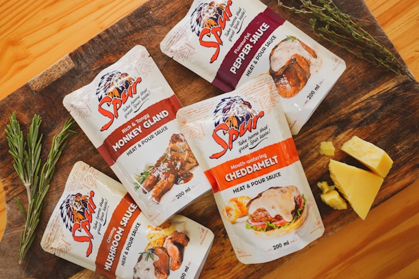 Five packets of Spur heat and pour sauces are arranged on a wooden surface. The flavors include Monkey Gland, Cheddamelt, Mushroom Sauce, and Pepper Sauce. Fresh sprigs of rosemary and thyme are placed beside the pouches, and there are chunks of cheese next to the Cheddamelt packet. The packaging is visually appealing, featuring images of the respective dishes the sauces can be used on.