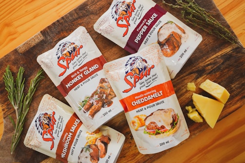 Five packets of Spur heat and pour sauces are arranged on a wooden surface. The flavors include Monkey Gland, Cheddamelt, Mushroom Sauce, and Pepper Sauce. Fresh sprigs of rosemary and thyme are placed beside the pouches, and there are chunks of cheese next to the Cheddamelt packet. The packaging is visually appealing, featuring images of the respective dishes the sauces can be used on.