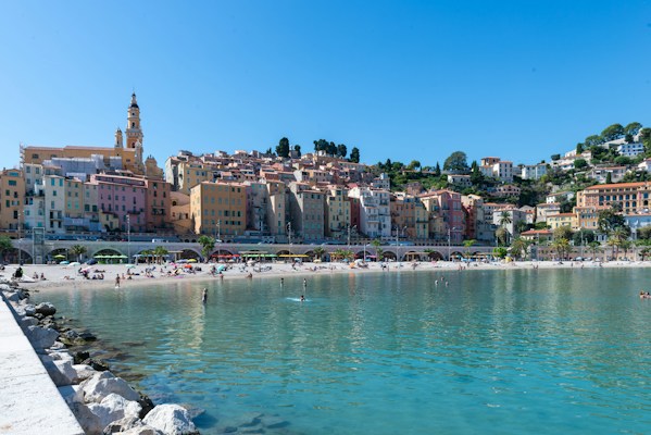 A picturesque coastal town with pastel-colored buildings stacked on a hillside overlooking a sandy beach. The clear blue sky complements the turquoise water, while people relax and enjoy the beach. The architecture features a prominent church with a tall bell tower, enhancing the charming Mediterranean vibe.