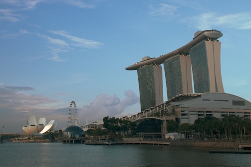 A panoramic view of Singapore's Marina Bay area, featuring the Marina Bay Sands integrated resort with its iconic three towers and boat-shaped structure on top. The ArtScience Museum with its lotus-inspired design is visible to the left, along with the Singapore Flyer ferris wheel. The sky is mostly clear with some clouds, and the water in the foreground reflects the buildings.