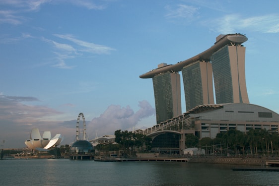A panoramic view of Singapore's Marina Bay area, featuring the Marina Bay Sands integrated resort with its iconic three towers and boat-shaped structure on top. The ArtScience Museum with its lotus-inspired design is visible to the left, along with the Singapore Flyer ferris wheel. The sky is mostly clear with some clouds, and the water in the foreground reflects the buildings.