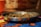 A metal bowl filled with ring-shaped dog food pellets sits on a colorful tablecloth. The background is blurred, featuring warm tones.