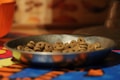 A metal bowl filled with ring-shaped dog food pellets sits on a colorful tablecloth. The background is blurred, featuring warm tones.