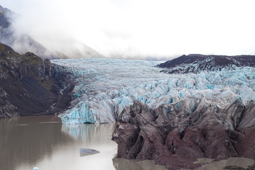 A vast glacier spreads across a mountainous landscape, with a mix of white and light blue hues indicating its icy terrain. Dark rocky mountains flank the glacier, contrasting sharply against the ice. In the foreground, a calm body of water reflects the glacier's edge, while misty clouds hover above, contributing to the dramatic scenery.