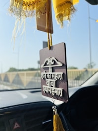 A hanging car ornament featuring a sign with text in a South Asian language, adorned with tassels. The background shows blurred vehicle interiors and a view of a road with barriers.