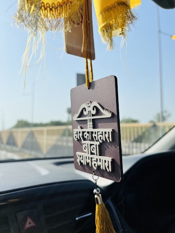 A hanging car ornament featuring a sign with text in a South Asian language, adorned with tassels. The background shows blurred vehicle interiors and a view of a road with barriers.