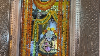 A beautifully adorned religious shrine featuring a deity figure surrounded by vibrant garlands of marigold flowers in shades of orange and yellow. The deity is intricately decorated with colorful ornaments and jewelry, set against an ornate backdrop with detailed metallic patterns. A figure clad in white is positioned behind, possibly a caretaker or spiritual guardian, adding a serene touch to the setting.