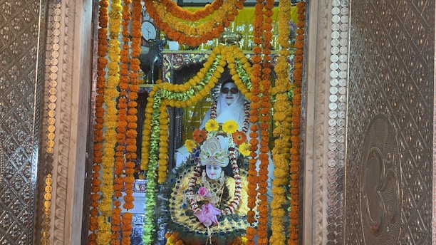 A beautifully adorned religious shrine featuring a deity figure surrounded by vibrant garlands of marigold flowers in shades of orange and yellow. The deity is intricately decorated with colorful ornaments and jewelry, set against an ornate backdrop with detailed metallic patterns. A figure clad in white is positioned behind, possibly a caretaker or spiritual guardian, adding a serene touch to the setting.