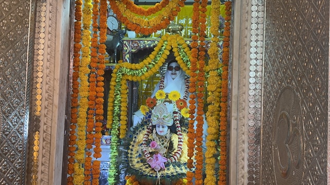 A beautifully adorned religious shrine featuring a deity figure surrounded by vibrant garlands of marigold flowers in shades of orange and yellow. The deity is intricately decorated with colorful ornaments and jewelry, set against an ornate backdrop with detailed metallic patterns. A figure clad in white is positioned behind, possibly a caretaker or spiritual guardian, adding a serene touch to the setting.