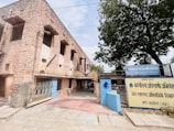 A stone building with a rustic appearance is situated on the left side, featuring multiple windows and a gated entrance. The foreground shows a paved walkway leading to the entrance, and a large tree is visible on the right. Signage in front of the building is in Hindi.