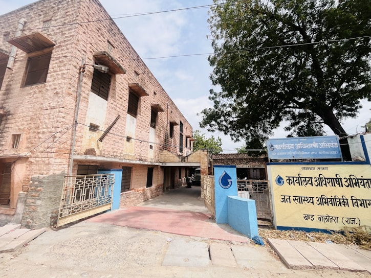 A stone building with a rustic appearance is situated on the left side, featuring multiple windows and a gated entrance. The foreground shows a paved walkway leading to the entrance, and a large tree is visible on the right. Signage in front of the building is in Hindi.