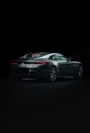 A sleek, modern sports car is displayed in a dimly lit setting. The vehicle has a shiny, reflective surface with elegant contours and sharp design lines. The rear and side profile highlight its aerodynamic shape, and the taillights emit a subtle red glow.