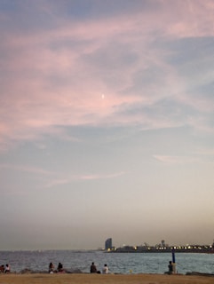A coastal scene during sunset with a sandy beach in the foreground where several people are sitting and enjoying the view. The calm sea meets the horizon beneath a sky painted with soft hues of pink and blue, and a crescent moon is faintly visible. In the distance, city lights and a silhouette of a tall, uniquely shaped building are seen along the shore.