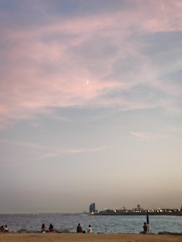 A coastal scene during sunset with a sandy beach in the foreground where several people are sitting and enjoying the view. The calm sea meets the horizon beneath a sky painted with soft hues of pink and blue, and a crescent moon is faintly visible. In the distance, city lights and a silhouette of a tall, uniquely shaped building are seen along the shore.