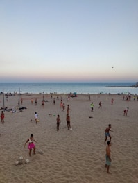 A beach scene with numerous people engaging in various ball games on the sand. The ocean is visible in the background, and the sky displays a gradient from blue to a pinkish hue near the horizon. Some individuals are standing, others are playing, and a few are sitting on the ground, enjoying the outdoor activity as the day begins to transition into evening.