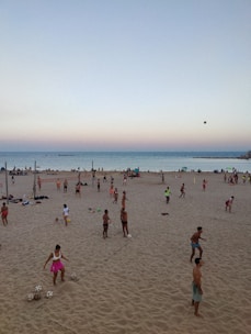 A beach scene with numerous people engaging in various ball games on the sand. The ocean is visible in the background, and the sky displays a gradient from blue to a pinkish hue near the horizon. Some individuals are standing, others are playing, and a few are sitting on the ground, enjoying the outdoor activity as the day begins to transition into evening.