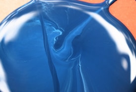 A smooth, glossy layer of blue liquid appears to be freshly poured, creating a swirling design with highlights and shadows. The surface texture suggests a viscous, flowing substance.
