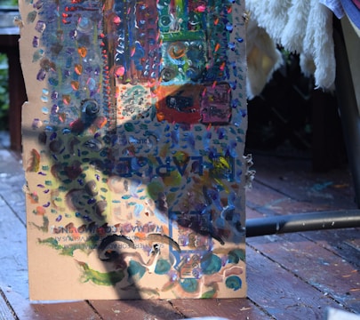 A piece of cardboard with colorful abstract paint strokes in various shades, including blues, greens, oranges, and purples. The background shows parts of a wooden deck and a fluffy white fabric draped over a chair.