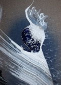 An abstract artwork featuring a blend of textured brushstrokes in white and dark blue, with a central splash of white paint against a dark background. The composition includes sweeping arcs and lines, leading to a sense of motion and fluidity.