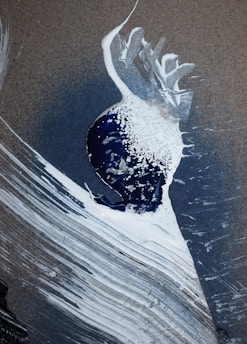 An abstract artwork featuring a blend of textured brushstrokes in white and dark blue, with a central splash of white paint against a dark background. The composition includes sweeping arcs and lines, leading to a sense of motion and fluidity.