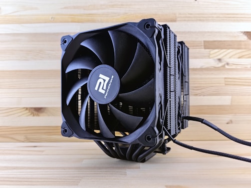 A close-up view of a black computer CPU cooler placed on a wooden surface. The cooler features prominent fan blades and heat dissipation fins. The branding is visible on the center hub of the fan, and power cables extend from the base of the cooler.