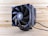 A close-up view of a black computer CPU cooler placed on a wooden surface. The cooler features prominent fan blades and heat dissipation fins. The branding is visible on the center hub of the fan, and power cables extend from the base of the cooler.