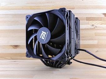 A close-up view of a black computer CPU cooler placed on a wooden surface. The cooler features prominent fan blades and heat dissipation fins. The branding is visible on the center hub of the fan, and power cables extend from the base of the cooler.
