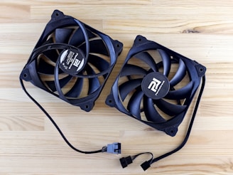 Two black computer cooling fans placed on a wooden surface. Each fan has a circular hub at the center with a logo and text. The fans have several blades and are connected by cables with connectors at the ends.