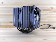 A large CPU cooler with a dual-tower heatsink design, equipped with two black cooling fans and multiple heat pipes. The background consists of a light wooden surface, enhancing the contrast with the dark color of the cooler.