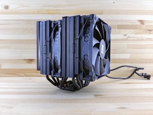 A large CPU cooler with a dual-tower heatsink design, equipped with two black cooling fans and multiple heat pipes. The background consists of a light wooden surface, enhancing the contrast with the dark color of the cooler.