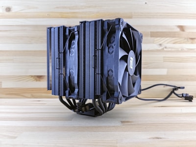 A large CPU cooler with a dual-tower heatsink design, equipped with two black cooling fans and multiple heat pipes. The background consists of a light wooden surface, enhancing the contrast with the dark color of the cooler.