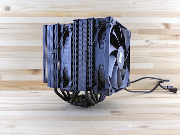 A large CPU cooler with a dual-tower heatsink design, equipped with two black cooling fans and multiple heat pipes. The background consists of a light wooden surface, enhancing the contrast with the dark color of the cooler.