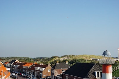 A coastal town with several houses featuring red roofs and a lighthouse with red and white stripes. There’s a small hill with grassy vegetation in the background and a clear blue sky.